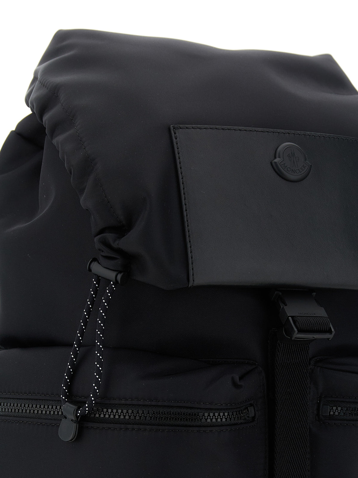 Close-up of a black backpack showing the top flap, a front pocket with a circular logo tab, and a zipper closure against a white background.