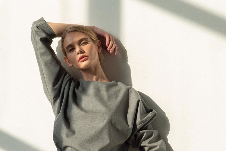 A woman models a matching two-piece gray set consisting of a loose long-sleeve top and wide-leg trousers, styled with a black leather belt featuring a gold buckle, set against a white wall with dramatic shadows.